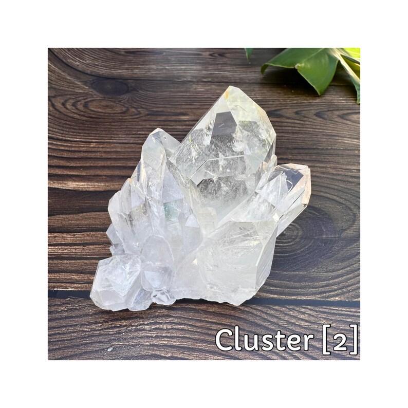 May include: A clear quartz crystal cluster with multiple points. The crystals are translucent and have a natural, rough texture. The cluster is labeled "Cluster [2]" in white text. The background is a dark wood surface.