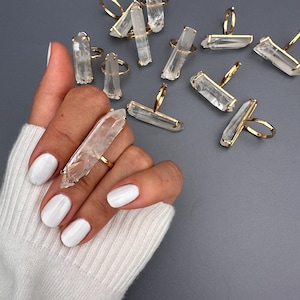 May include: A hand displaying a gold ring featuring a large, clear quartz crystal. Other rings with quartz crystals are on a grey surface. The rings have a simple design, highlighting the natural beauty of the crystals. The crystals are approximately 5 cm in length.