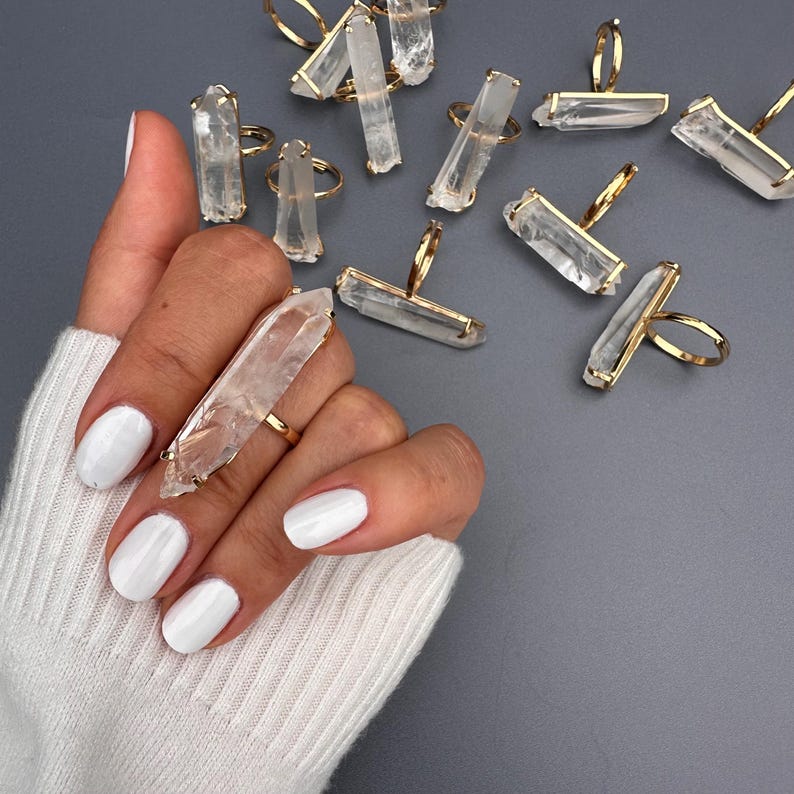 May include: A hand displaying a gold ring featuring a large, clear quartz crystal. Other rings with quartz crystals are on a grey surface. The rings have a simple design, highlighting the natural beauty of the crystals. The crystals are approximately 5 cm in length.