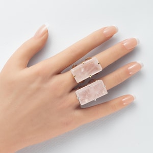 May include: Two rectangular rings featuring large, pale pink rose quartz stones set in metal bands. The top ring has a gold-toned setting, while the bottom ring features a silver-toned setting, both worn on adjacent fingers.