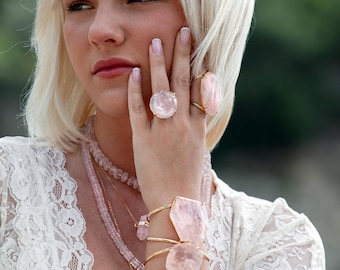 Puede incluir: Dos anillos dorados con grandes piedras de cuarzo rosa redondas y ovaladas, un brazalete dorado con dos grandes detalles de cuarzo rosa angulares y un collar de varias capas con cuentas rosas facetadas y un pequeño colgante de cuarzo rosa.