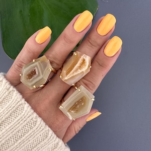 May include: Three gold rings featuring beige agate stones. The stones have a natural, raw appearance with a crystal-like centre. The rings are stacked on a hand with yellow nail polish.