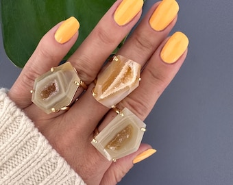 Puede incluir: Tres anillos de oro con piedras de ágata beige. Las piedras tienen un aspecto natural y crudo con un centro cristalino. Los anillos están apilados en una mano con esmalte de uñas amarillo.