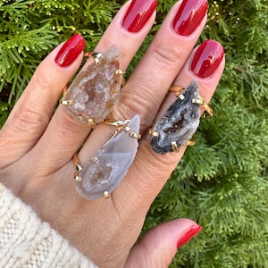 May include: Three gemstone rings with gold-colored bands. The gemstones are teardrop-shaped, with colors ranging from light gray to brown and black. The rings are displayed on a hand with red nail polish, against a backdrop of green foliage.