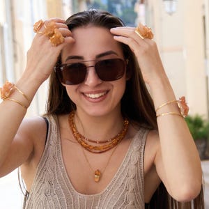 May include: A woman wearing sunglasses and a collection of orange-toned jewelry. The jewelry includes rings, bracelets, and layered necklaces. The bracelets and rings feature a floral design. The necklaces have a pendant.