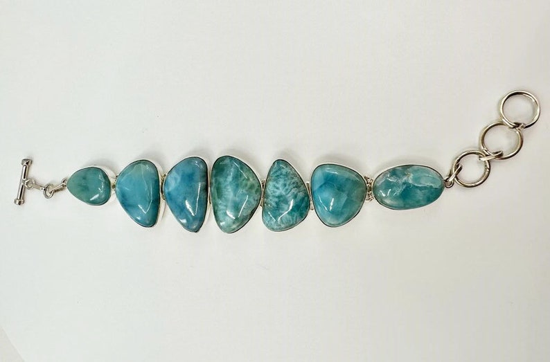 May include: A silver bracelet with a row of six light blue and green gemstone beads. The bracelet has a toggle clasp on one end and a ring clasp on the other.
