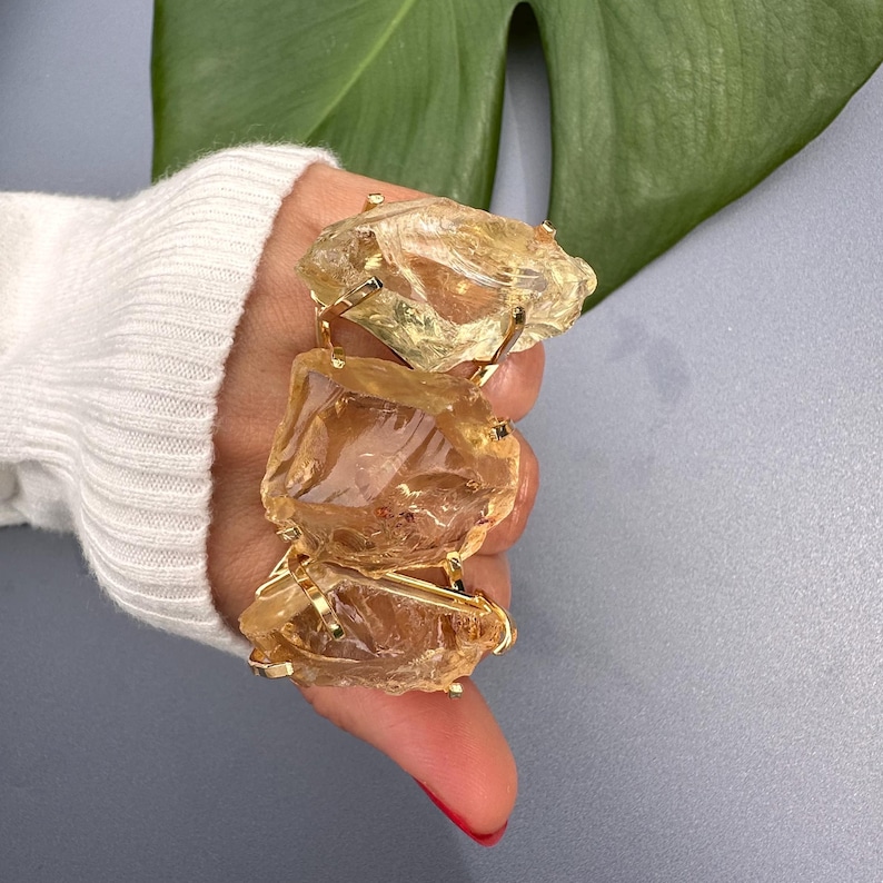May include: A gold-toned bracelet featuring three large, irregularly shaped, translucent yellow-brown gemstones. The stones are held together by delicate gold metal connectors. The bracelet is displayed against a neutral background.
