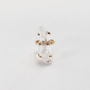 May include: A clear quartz crystal ring with a gold band and gold detailing. The crystal is vertically aligned and has a natural, textured appearance. The ring is set against a white backdrop.