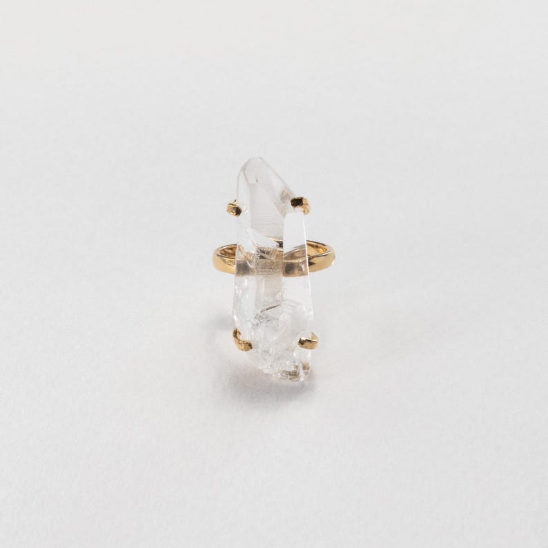 May include: A clear quartz crystal ring with a gold band and gold detailing. The crystal is vertically aligned and has a natural, textured appearance. The ring is set against a white backdrop.