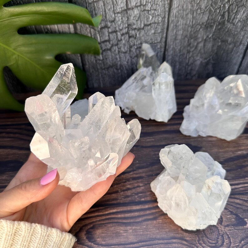 May include: Clear quartz crystal clusters. The image shows several clear quartz crystal clusters of varying sizes. The crystals are translucent and have a natural, rough texture. The crystals are displayed on a dark wooden surface.