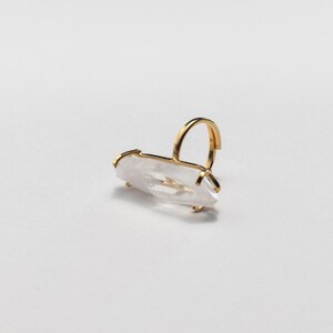 May include: A gold ring with a clear, elongated crystal. The crystal is held in place by a gold setting. The ring's band is a simple, curved design. The ring is set against a white background.