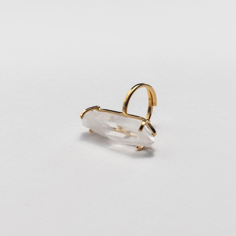 May include: A gold ring with a clear, elongated crystal. The crystal is held in place by a gold setting. The ring's band is a simple, curved design. The ring is set against a white background.