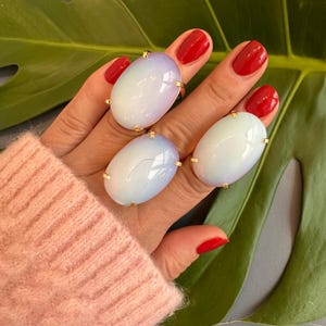 May include: Three oval-shaped rings with iridescent white and blue stones set in gold settings. The rings are worn on the fingers of a hand.