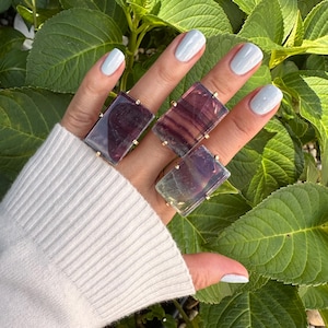 May include: Three rectangular gemstone rings with gold-colored settings are displayed on a hand. The gemstones are a mix of purple, green, and clear hues. The hand is wearing a white sweater and has light blue nail polish. Green leaves are in the background.