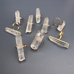 May include: Several gold rings, each adorned with a clear, elongated crystal. The crystals have a natural, raw appearance, with slight variations in shape and size. The rings are displayed on a grey surface, highlighting their unique design.