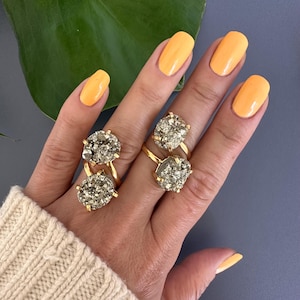 May include: Two gold rings featuring large, oval-shaped pyrite stones. The rings are worn on the index and middle fingers of a hand with yellow nail polish.