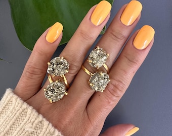 Puede incluir: Dos anillos de oro con grandes piedras ovaladas de pirita. Los anillos se llevan en el índice y el dedo medio de una mano con esmalte de uñas amarillo.