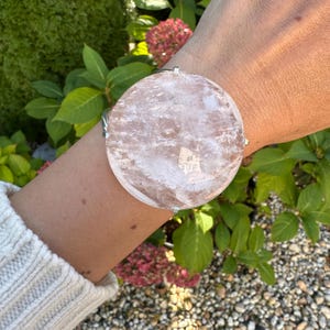 May include: A large, round, clear quartz bracelet with a silver-colored metal band. The quartz has a soft pink hue and is set against a background of green foliage and pink flowers.