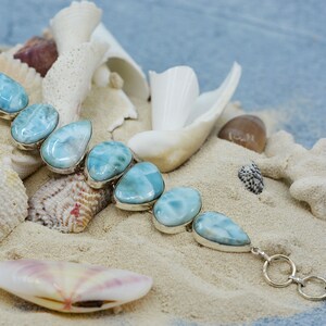 May include: A silver bracelet with a row of light blue teardrop-shaped stones. The bracelet is on a bed of white sand with seashells.