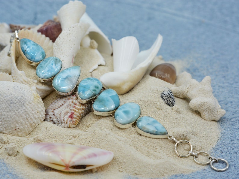 May include: A silver bracelet with a row of light blue teardrop-shaped stones. The bracelet is on a bed of white sand with seashells.