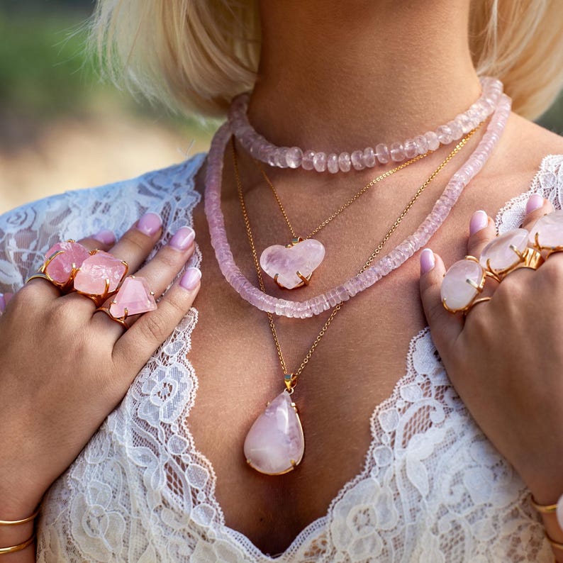 Puede incluir: Una colecci&oacute;n de joyas de cuarzo rosa, que incluye collares, anillos y pulseras. Los collares presentan colgantes en forma de coraz&oacute;n y de l&aacute;grima, as&iacute; como hileras de cuentas. Los anillos muestran piedras grandes en bruto engastadas en metal dorado. Las joyas son de color rosa suave.