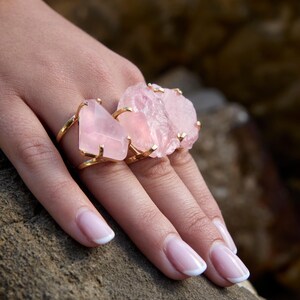Puede incluir: Dos anillos dorados con grandes piedras de cuarzo rosa claro. Uno tiene una piedra triangular, el otro una piedra rugosa e irregular. Los anillos se muestran en una mano con u&ntilde;as cuidadas.