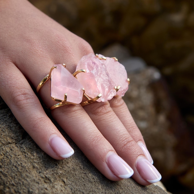 Puede incluir: Dos anillos dorados con grandes piedras de cuarzo rosa claro. Uno tiene una piedra triangular, el otro una piedra rugosa e irregular. Los anillos se muestran en una mano con u&ntilde;as cuidadas.