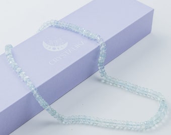 May include: A light blue aquamarine gemstone necklace with a delicate, faceted bead design. The necklace is displayed on a light purple box with the text "CRYSTALLIKE" printed on it.