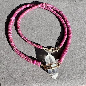 May include: A pink ruby bead necklace with a clear quartz crystal pendant. The necklace is made of small, round beads and has a gold clasp.