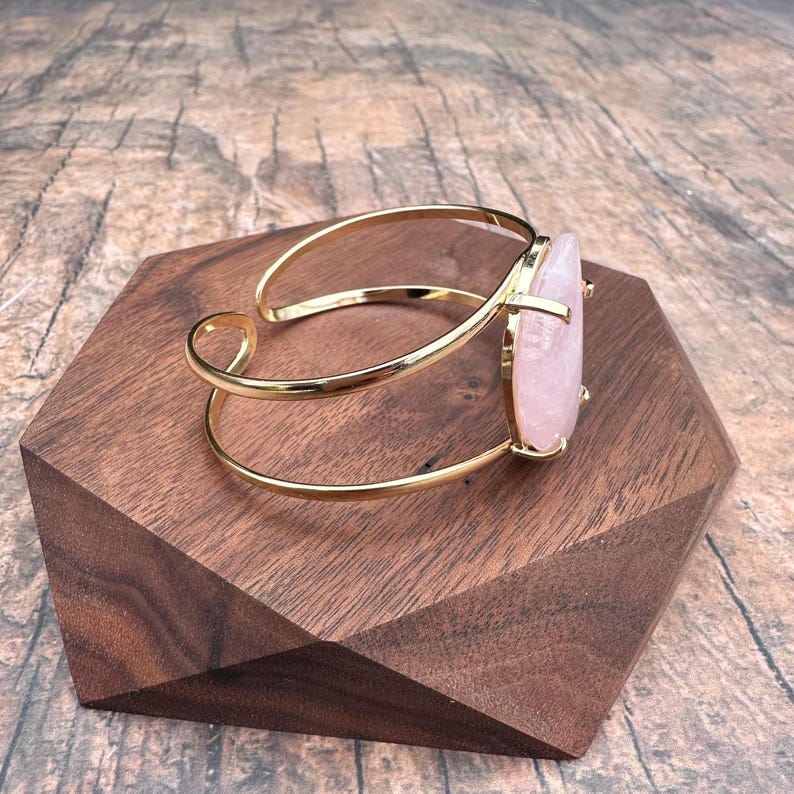 May include: A gold cuff bracelet with a large, oval-shaped rose quartz stone. The stone is a pale pink color and has a natural, irregular shape.