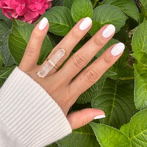 May include: A gold ring with a large, rectangular clear crystal. The ring is worn on a hand with white nail polish, set against green leaves and pink flowers. The crystal's shape is the main feature of the ring, which has a simple design.