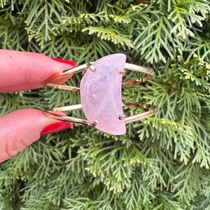 May include: A gold metal bangle bracelet with a large, half-moon shaped rose quartz stone.