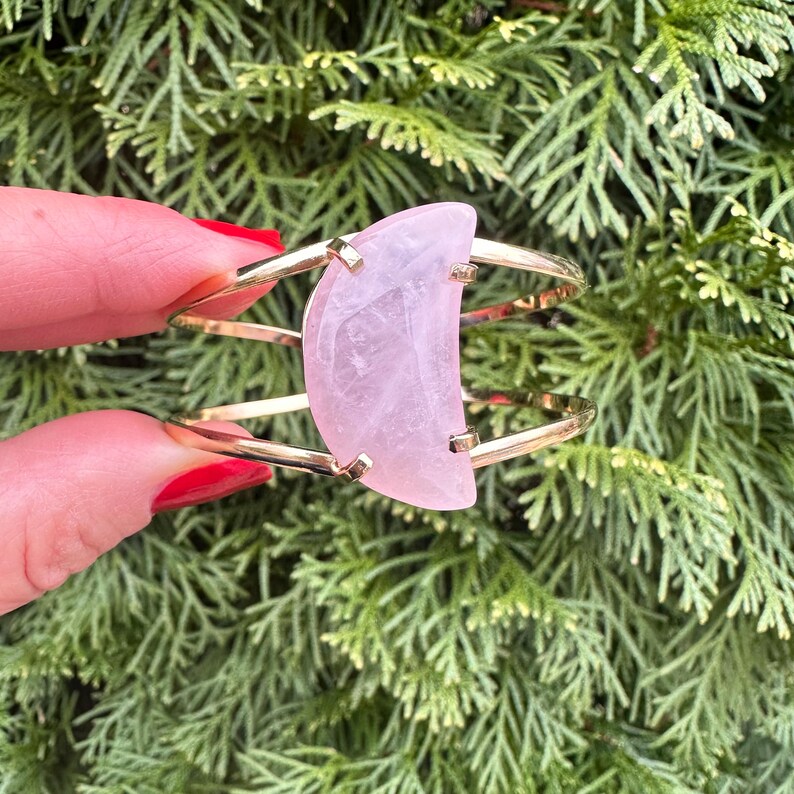 May include: A gold metal bangle bracelet with a large, half-moon shaped rose quartz stone.