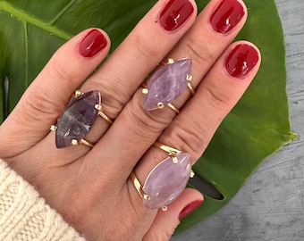 May include: Three gold rings, each featuring a large, oval amethyst gemstone, are shown on a hand. The amethyst stones display varying shades of purple. The rings are set against a green leaf and a grey background.