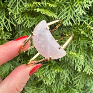 May include: A gold bangle bracelet with a large, pale pink, crescent-shaped rose quartz stone.