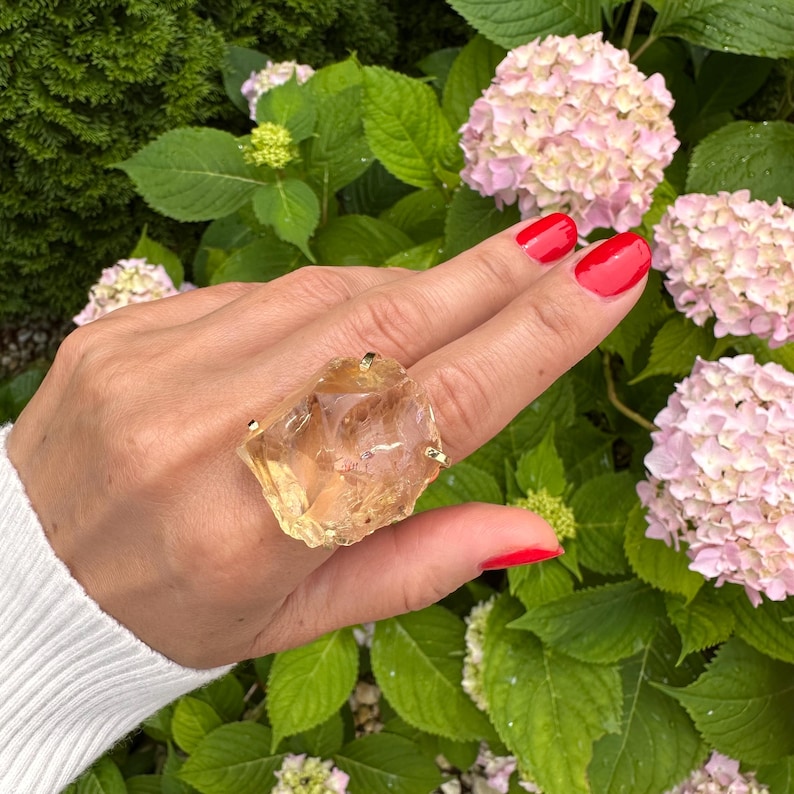 May include: A large, raw-cut citrine ring with gold prongs. The gemstone is a translucent amber color. The ring is displayed on a hand with red nail polish, set against a backdrop of green foliage and pink hydrangea flowers.