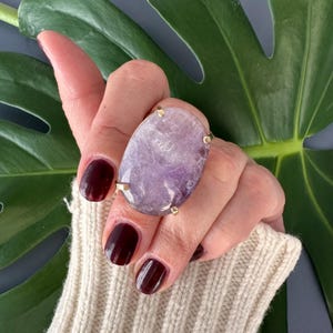 May include: A large oval amethyst ring with a gold setting. The ring is on a hand with dark red nail polish.