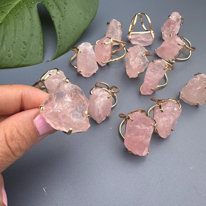 Puede incluir: Anillos de cuarzo rosa con engastes de metal dorado. Los anillos presentan piedras de cuarzo rosa en bruto, de forma irregular y de varios tama&ntilde;os. El fondo es de color gris s&oacute;lido, con algunas hojas verdes visibles.