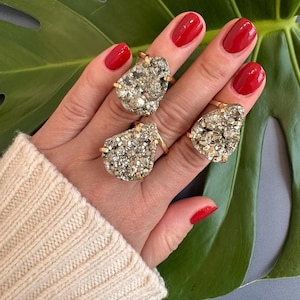 May include: Three gold rings featuring large, teardrop-shaped pyrite stones. The rings are worn on a hand with red nail polish.