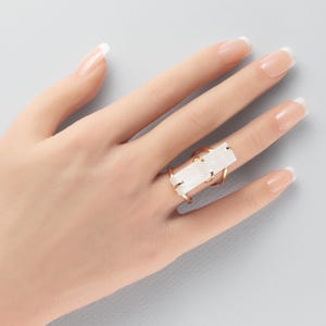 May include: A gold-toned ring featuring a long, rectangular, translucent white stone set horizontally. The metal band has a simple, open design that secures the stone with four small prongs at each corner, creating a clean and geometric look.
