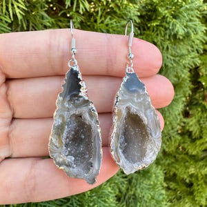 May include: A pair of silver earrings with a teardrop-shaped agate stone. The agate is a natural crystal with a gray and white color and a hollow center.