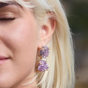 May include: Gold-framed amethyst earrings. Each earring features two clusters of purple crystals. The earrings have a gold-colored square frame. The amethyst crystals have a natural, rough texture. The earrings are a unique jewelry item.