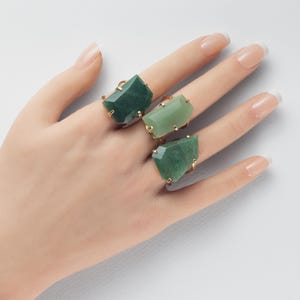 May include: Three rings featuring large, faceted, irregular-shaped green stones set in gold-toned metal bands. The stones vary in shade from deep forest green to a lighter, translucent mint green, each secured by small metal prongs on the sides.