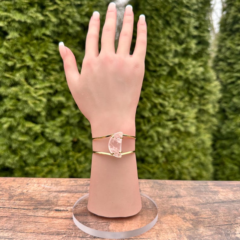 May include: A gold wire bracelet with a clear, half-moon shaped crystal. The bracelet is on a mannequin wrist.