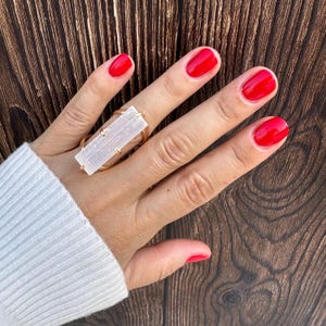 May include: A ring featuring a rectangular, translucent white selenite crystal bar set horizontally in a gold-toned metal band. The crystal has a fibrous, linear texture and is secured by small metal prongs at each end of the rectangular stone.