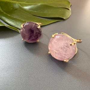 May include: Two gold rings with large round gemstone settings. The ring on the left has a purple amethyst stone. The ring on the right has a pink rose quartz stone.