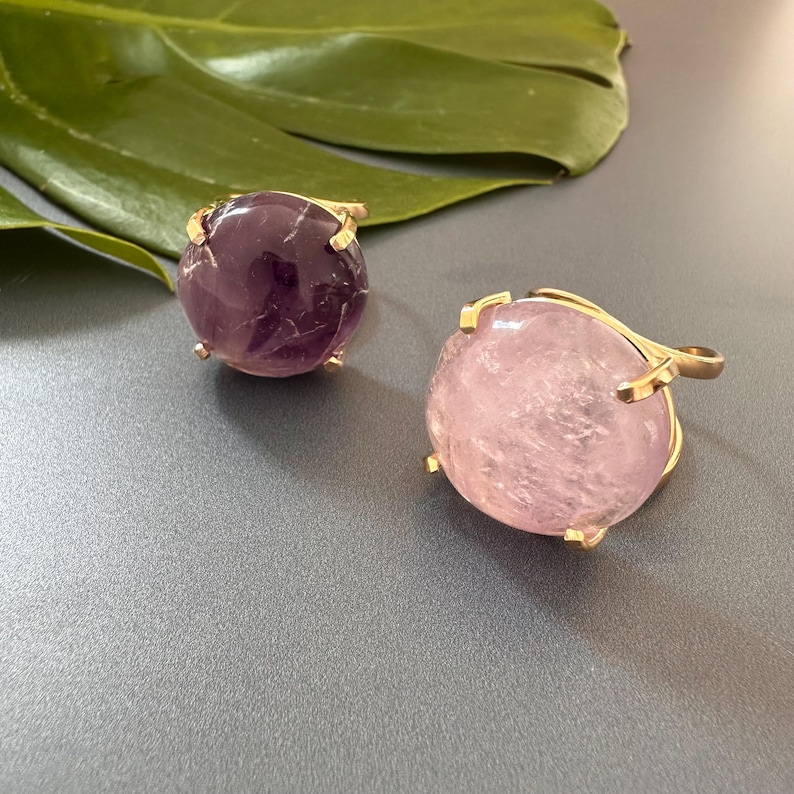 May include: Two gold rings with large round gemstone settings. The ring on the left has a purple amethyst stone. The ring on the right has a pink rose quartz stone.