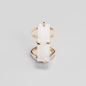 May include: A gold-toned ring featuring a vertical, rectangular, translucent white stone set in the centre. The metal band splits into two thin, curved sections that connect to the top and bottom of the stone, creating an open, geometric design.