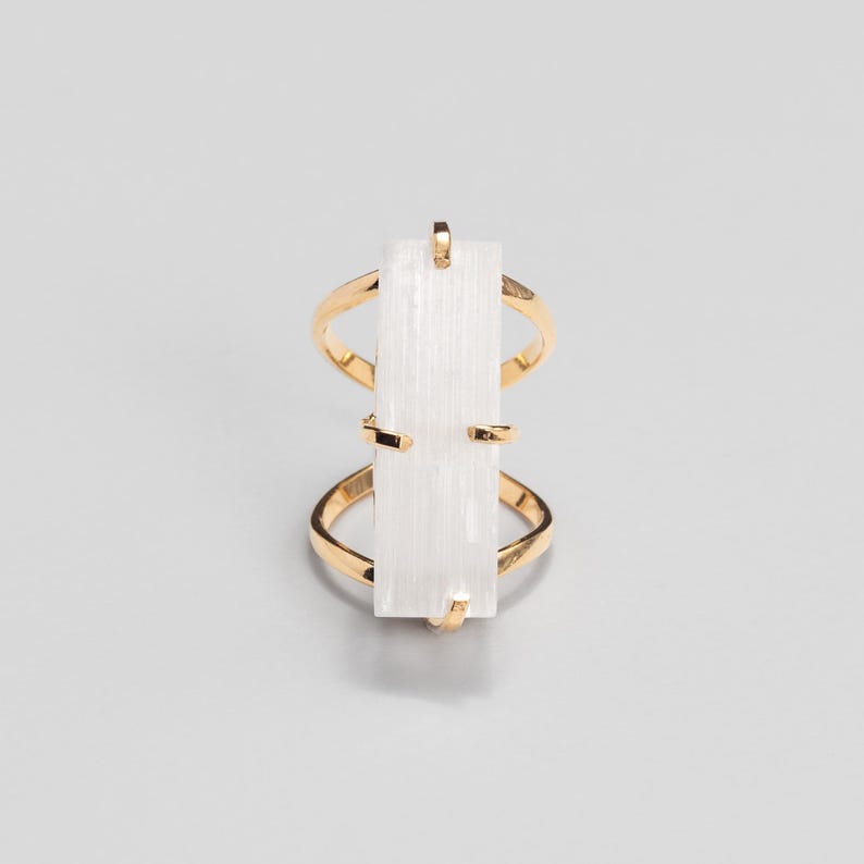 May include: A gold-toned ring featuring a vertical, rectangular, translucent white stone set in the centre. The metal band splits into two thin, curved sections that connect to the top and bottom of the stone, creating an open, geometric design.