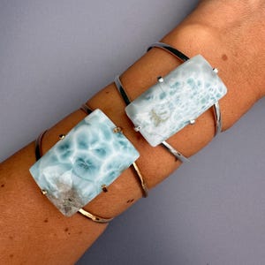 May include: Two silver and gold-toned cuff bracelets each feature a rectangular, light blue gemstone with white veining. The gemstones are mounted on the bracelets with small silver and gold-toned accents. The bracelets are worn on an arm against a gray background.